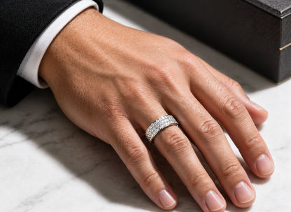 Close-up of a man's hand wearing a Mens Moissanite 3-Row Half Eternity Wedding Band 1.5CT | GRA with sparkling stones, resting on a marble surface next to a black box.